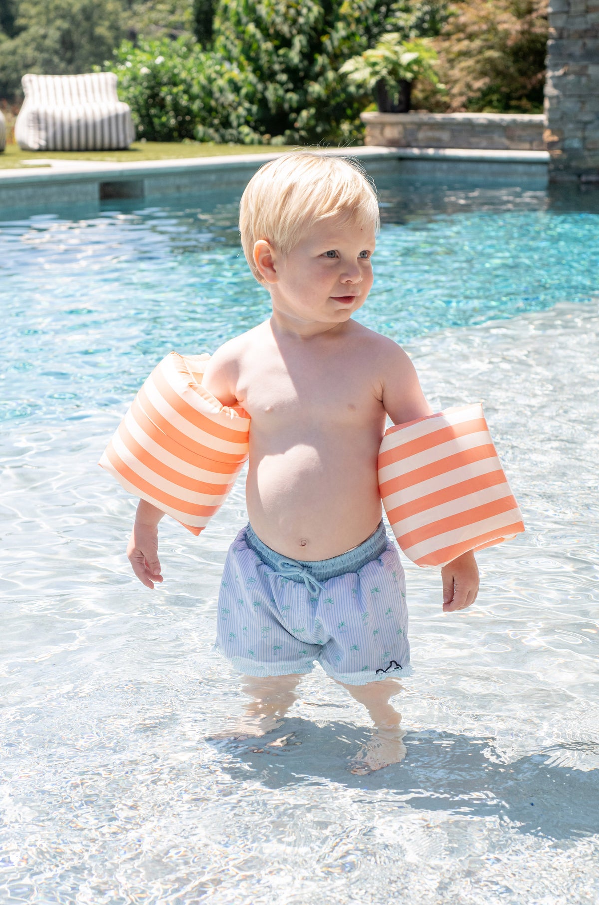 boy toddler in a pool wearing striped arm floats and boogie board swim shorts with palm trees and pinstripes