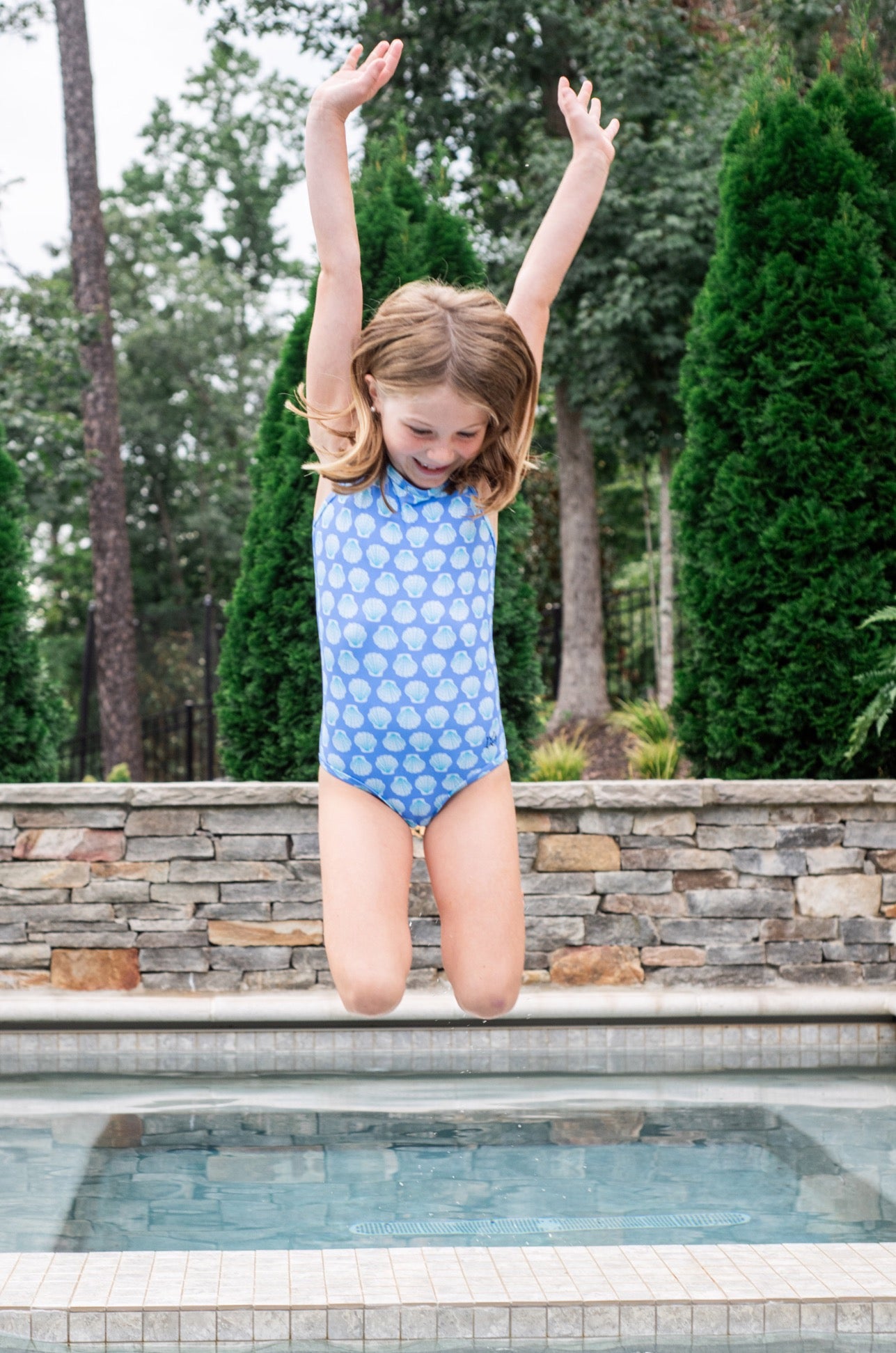 Young girl in a blue seashell print swimsuit jumping into a pool