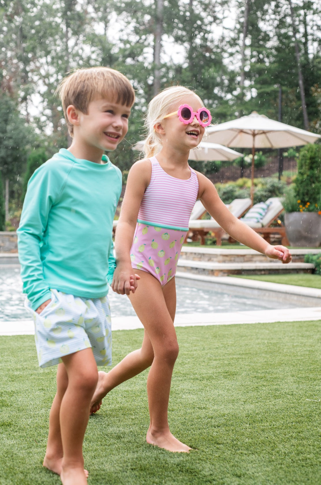Two children, a boy and a girl, standing on grass near a pool with trees in the background.