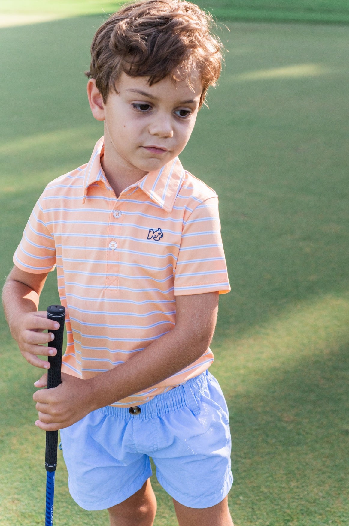 Young boy in an orange striped polo shirt and light blue shorts holding a golf club on a golf course.