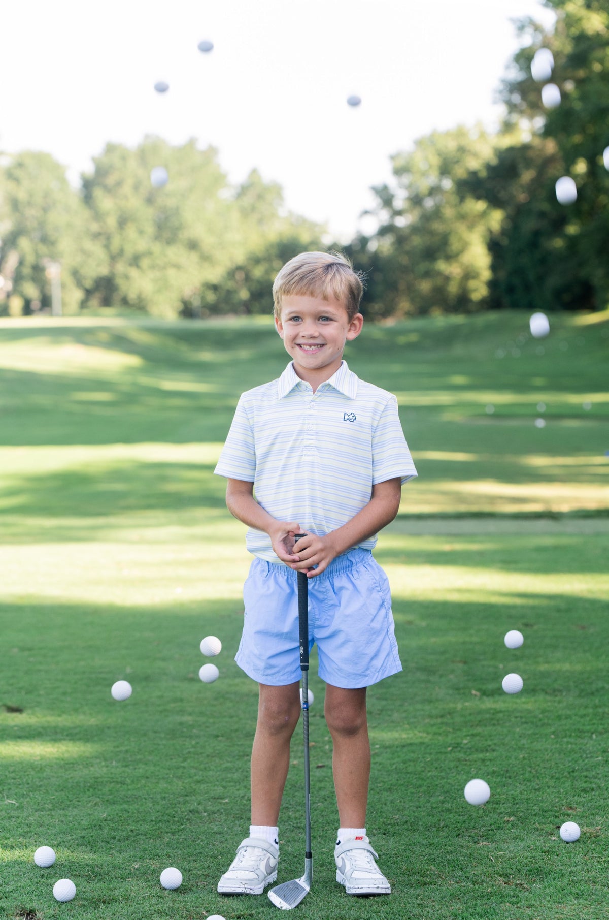Young boy on a golf course with golf balls around him wearing a yellow striped polo shirt