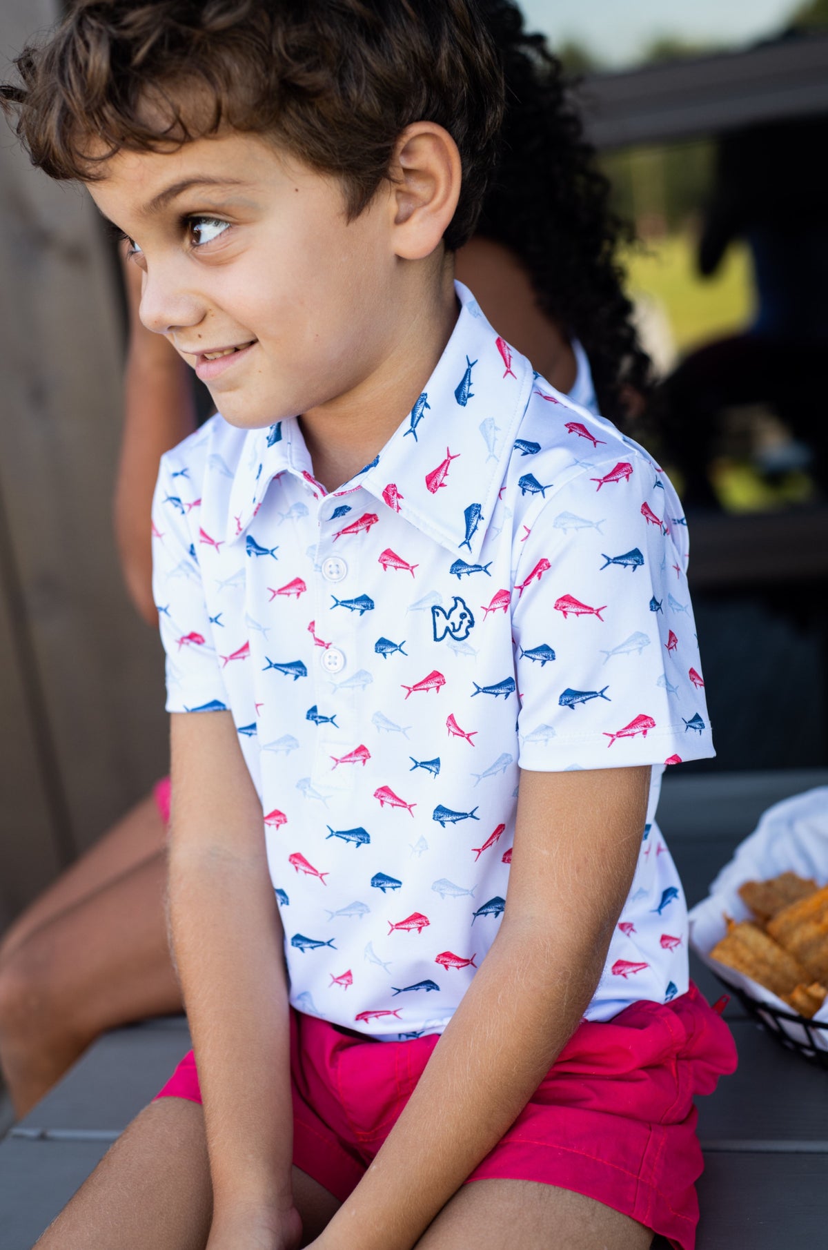 Child wearing a patterned quick-dry moisture wicking polo shirt and red shorts sitting outdoors.