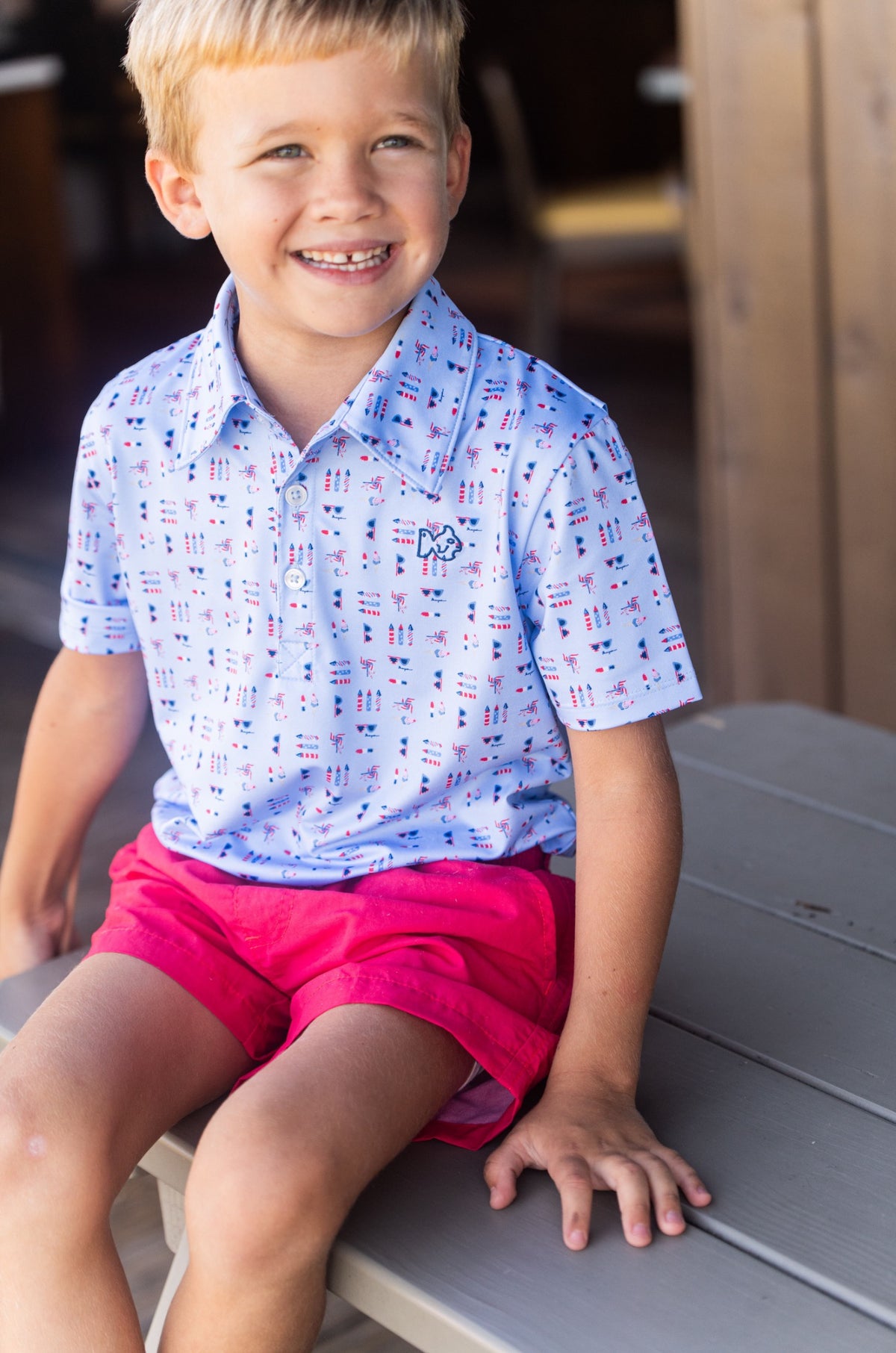 Young boy wearing a 4th of july patterned polo shirt  and red shorts sitting on a bench.