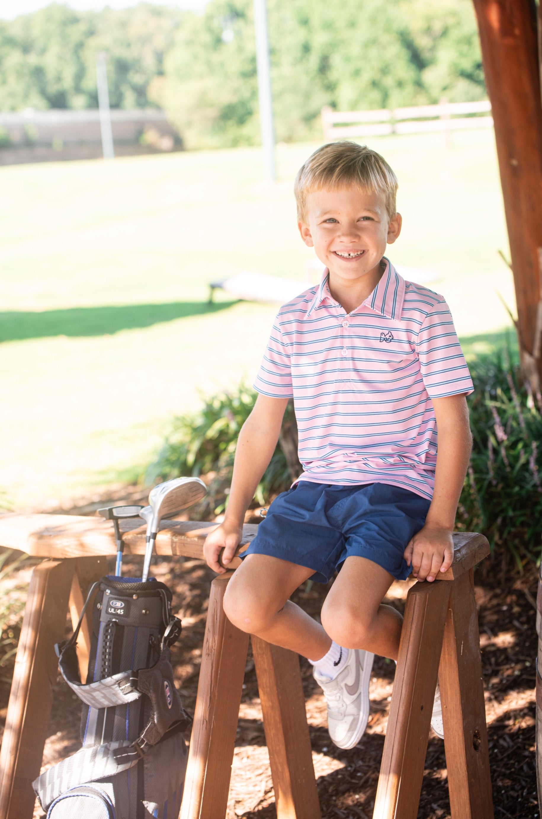 Young boy in a pink striped polo shirt sitting on a wooden stool outdoors with a golf club in the background.
