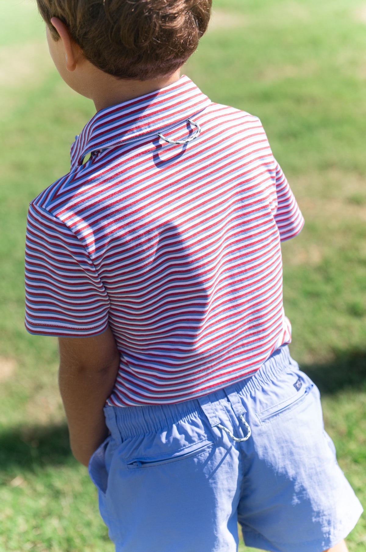 Child wearing a red and white striped polo shirt with blue shorts on a grassy background
