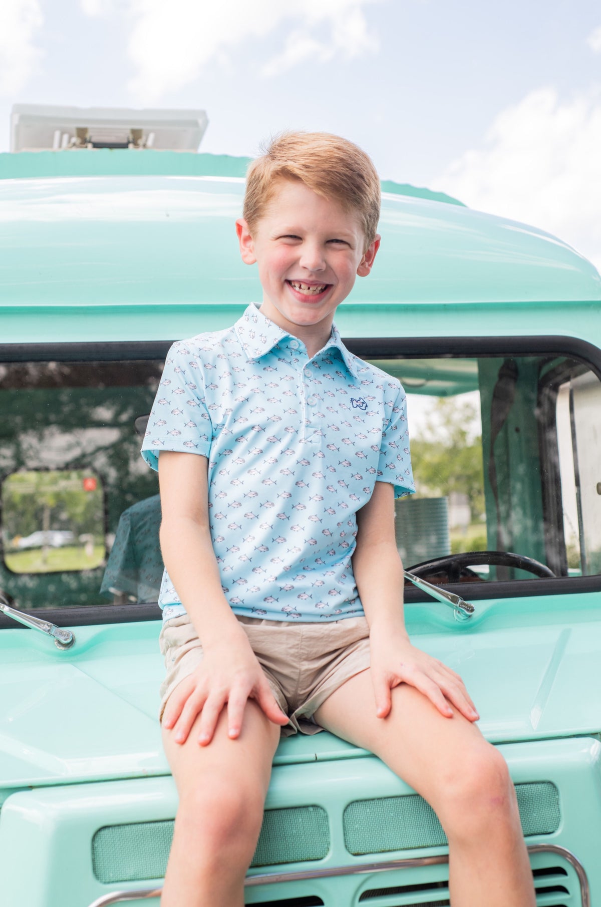Young boy sitting on a teal truck wearing activewear polo with small fish print designs