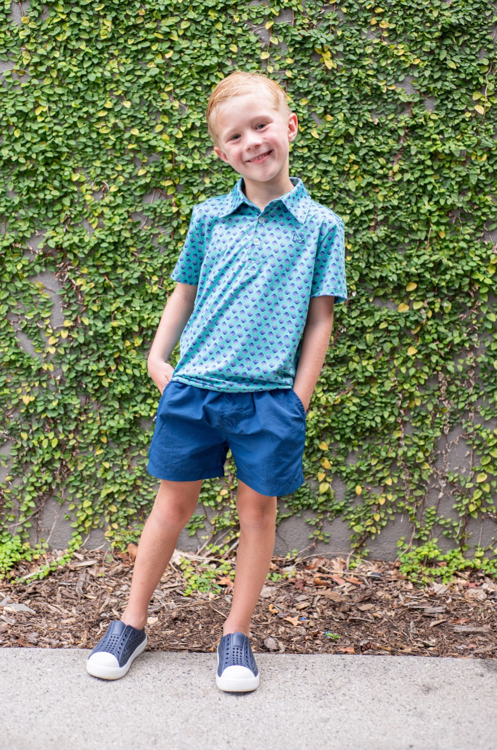 Young boy wearing a blue patterned shirt and shorts standing in front of a green ivy wall.