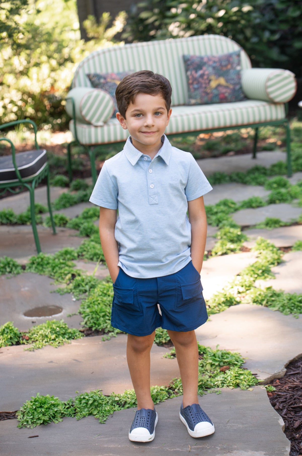 Young boy in a light blue cotton short-sleeve polo shirt and navy shorts standing outdoors on a patio with greenery.