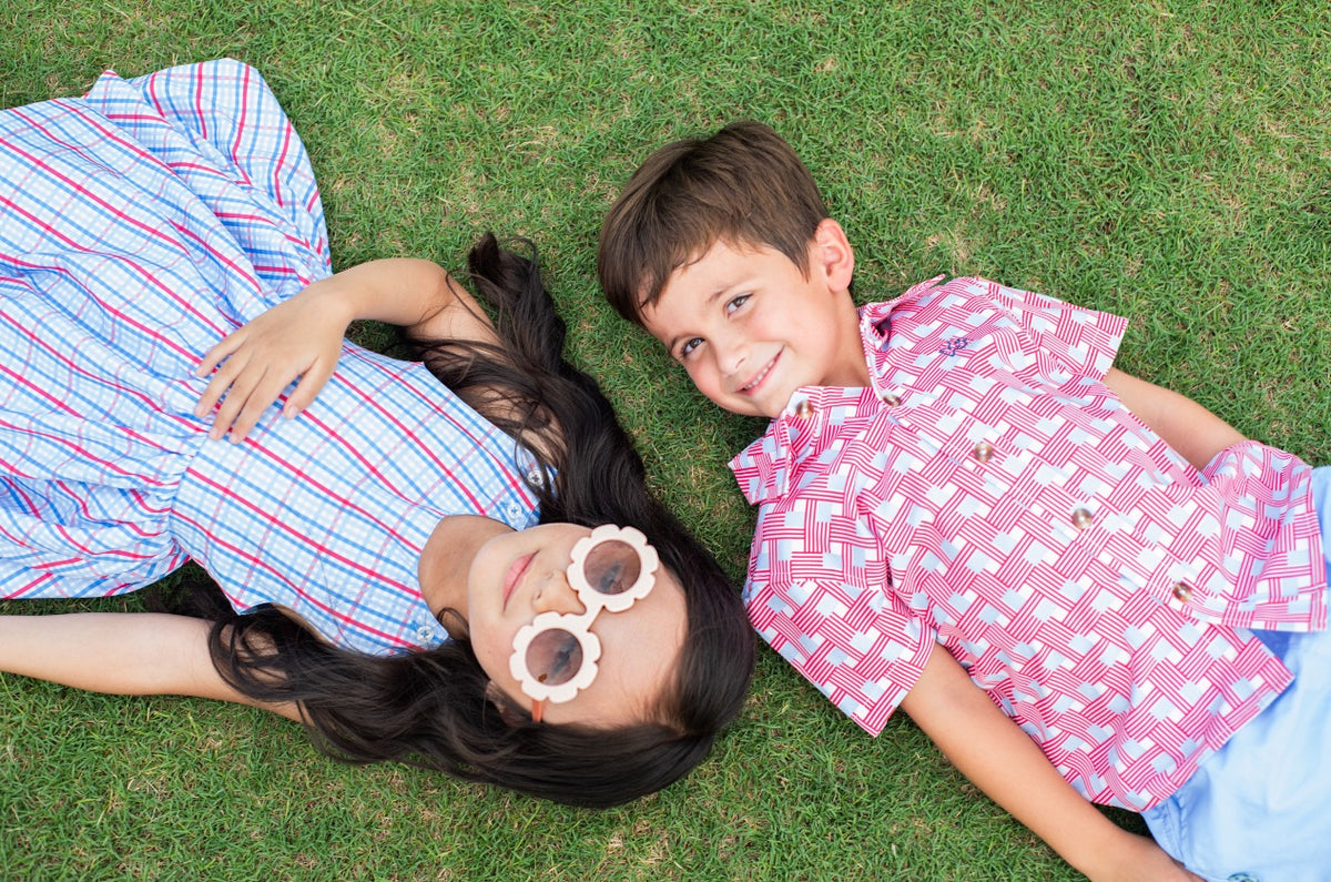 Two children lying on grass, with boy  wearing a red, white and blue checked woven button up shirt