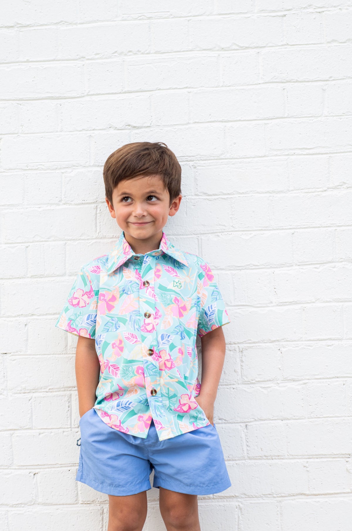 Child wearing a colorful floral shirt and blue shorts against a white brick wall.