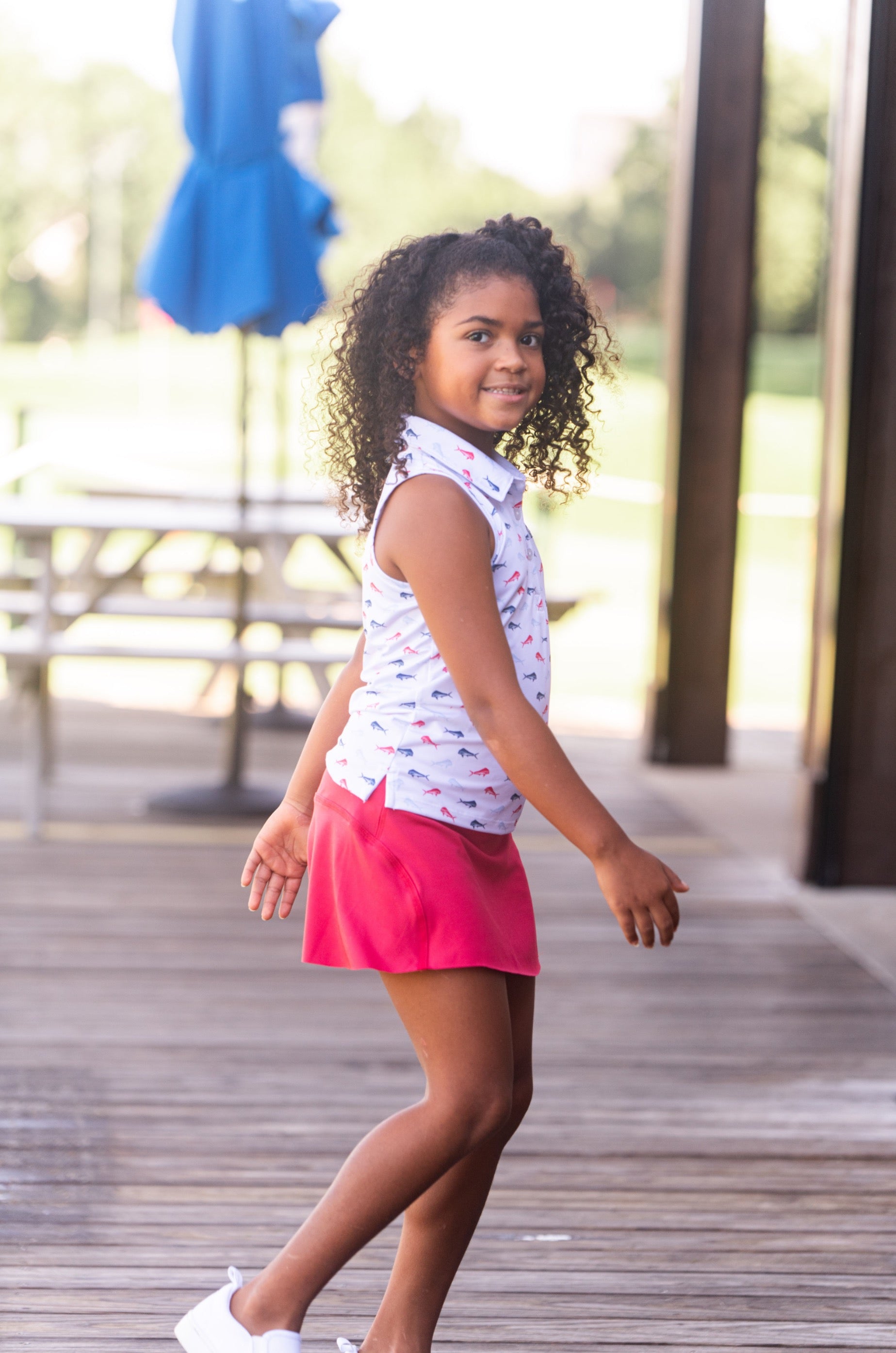 Young girl in a white sleeveless top and pink skirt walking outdoors on a wooden deck.