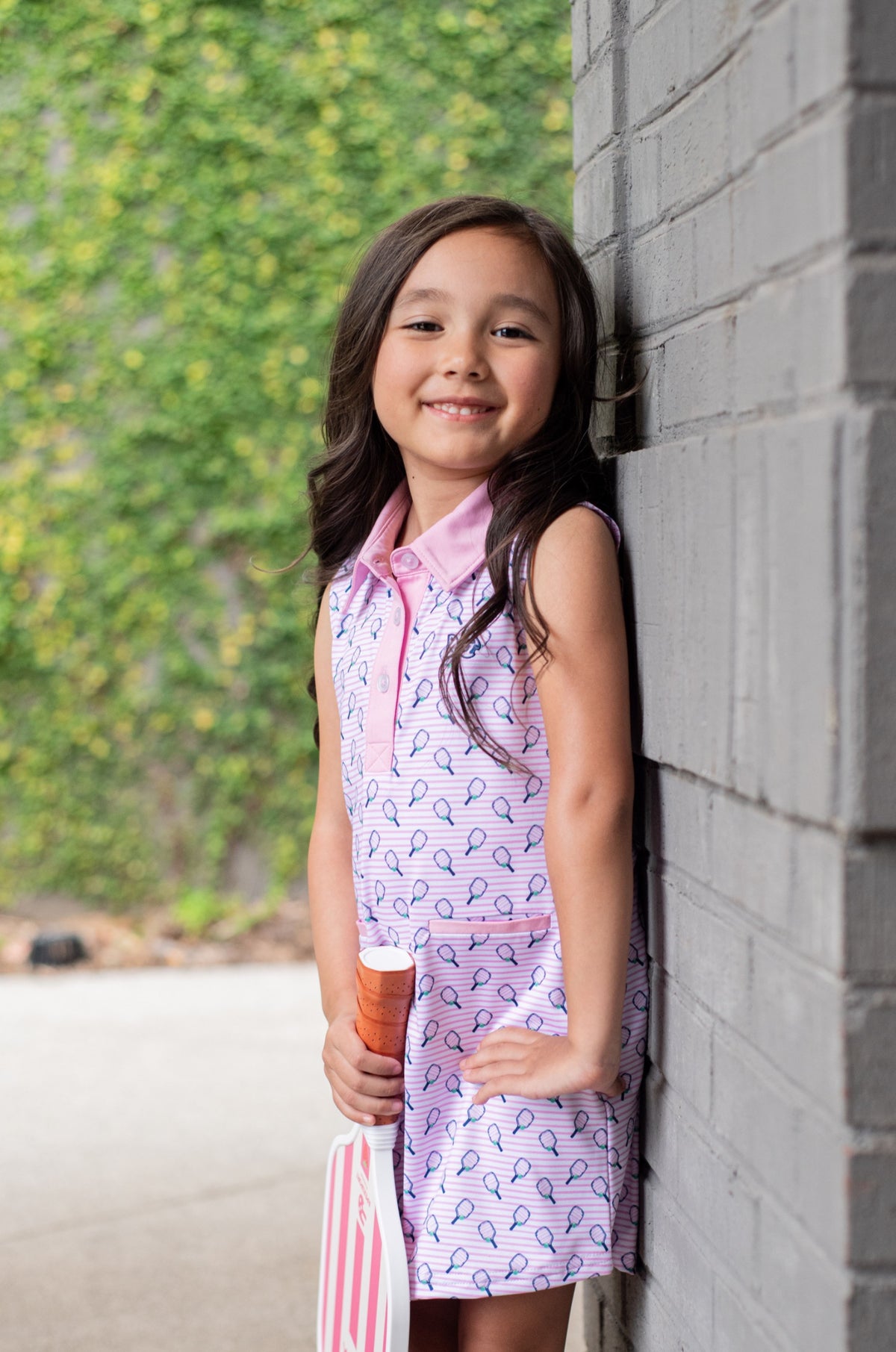 Young girl in a sleeveless polo dress leaning against a brick wall with greenery in the background