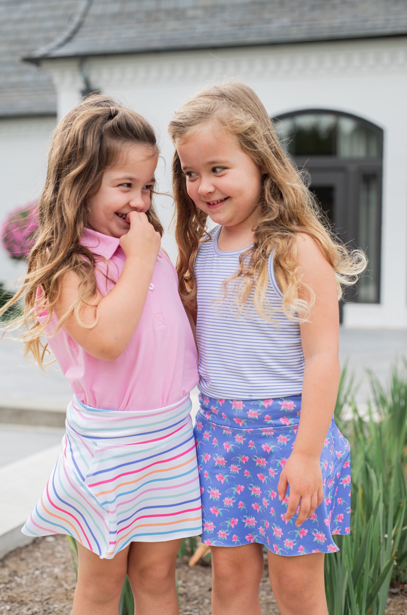 Two young girls standing together outdoors, wearing tennis skorts.