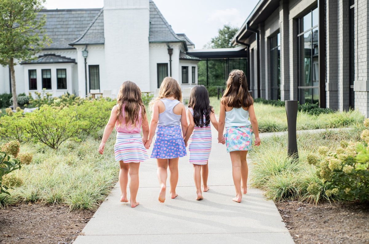 Four young girls in matching multi-color skorts walking on a path in a garden.