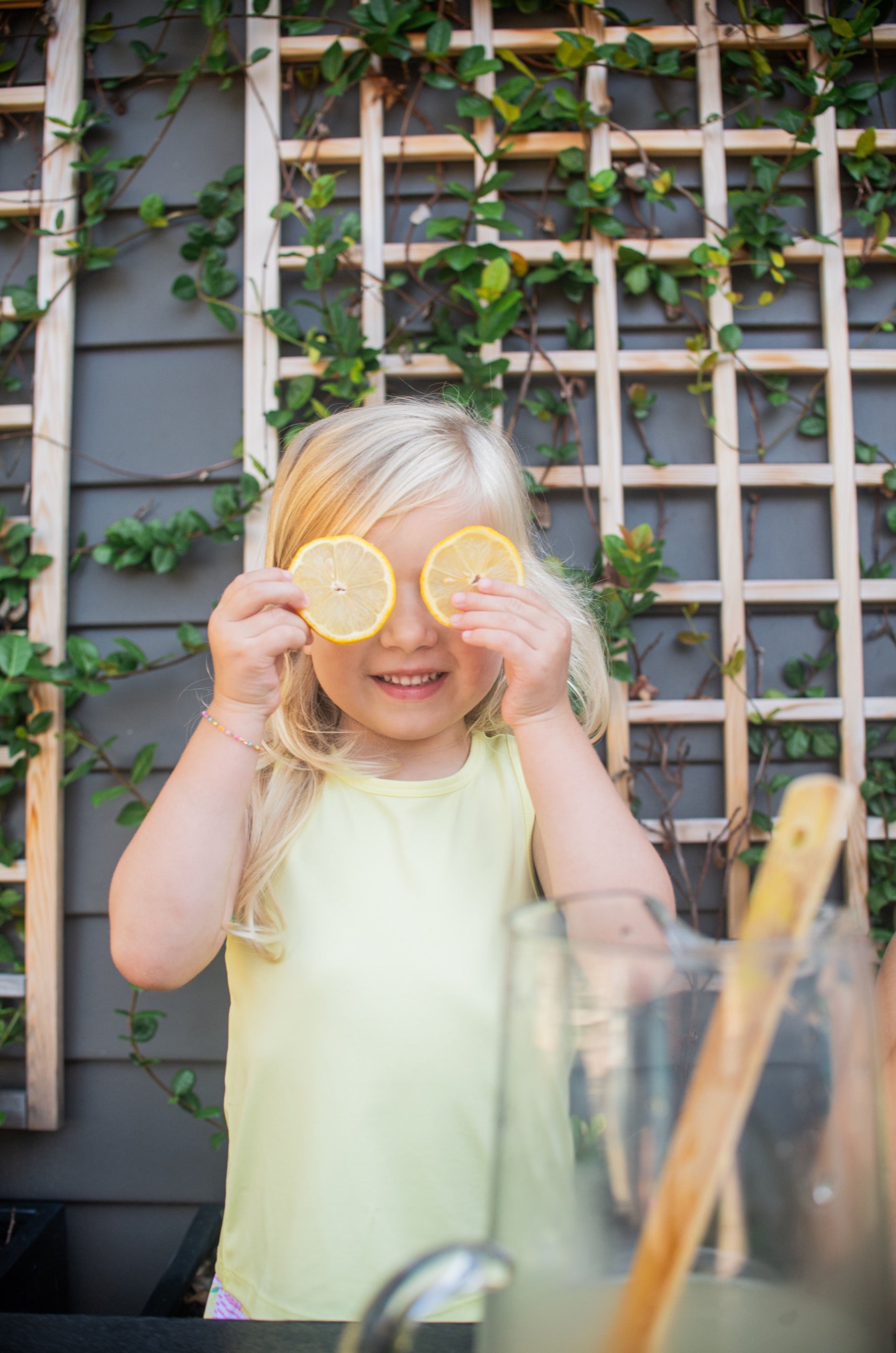 Young girl wearing yellow Tank Top and holding lemon slices over eyes with a garden background