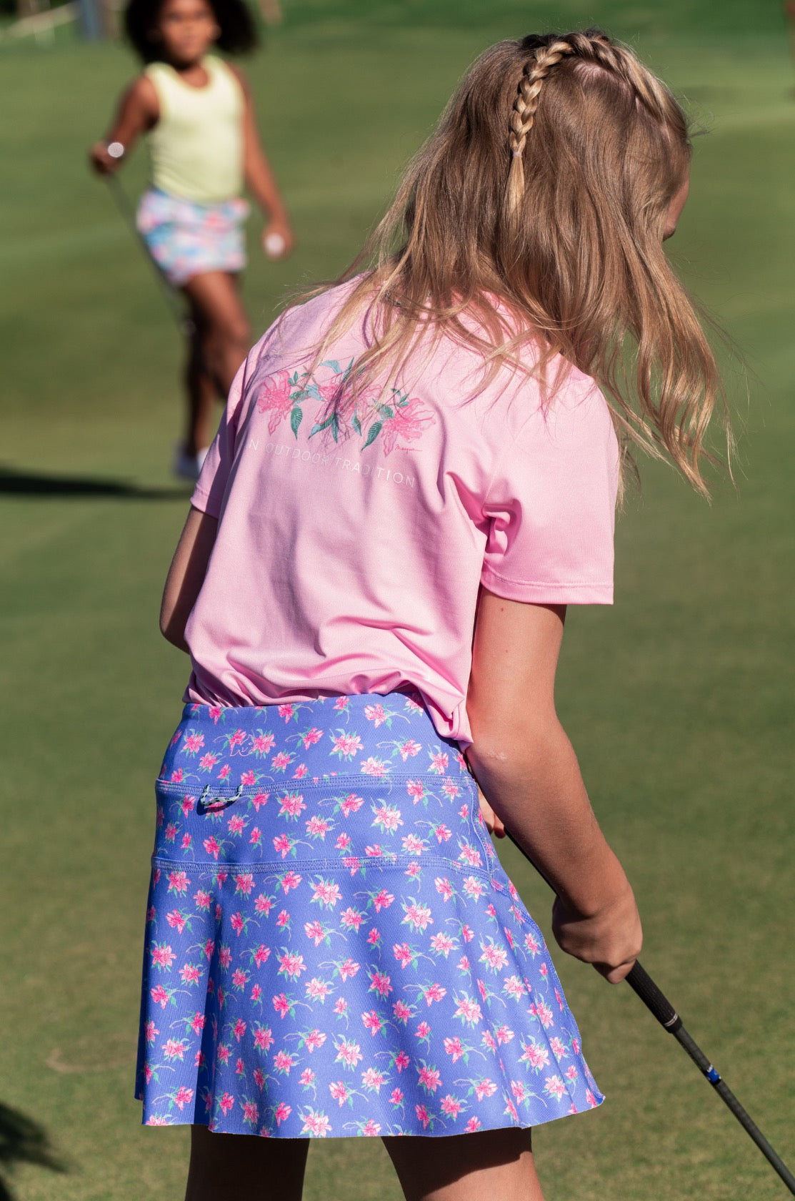 Girl in pink t-shirt and blue floral skirt holding a golf club on a grassy field.