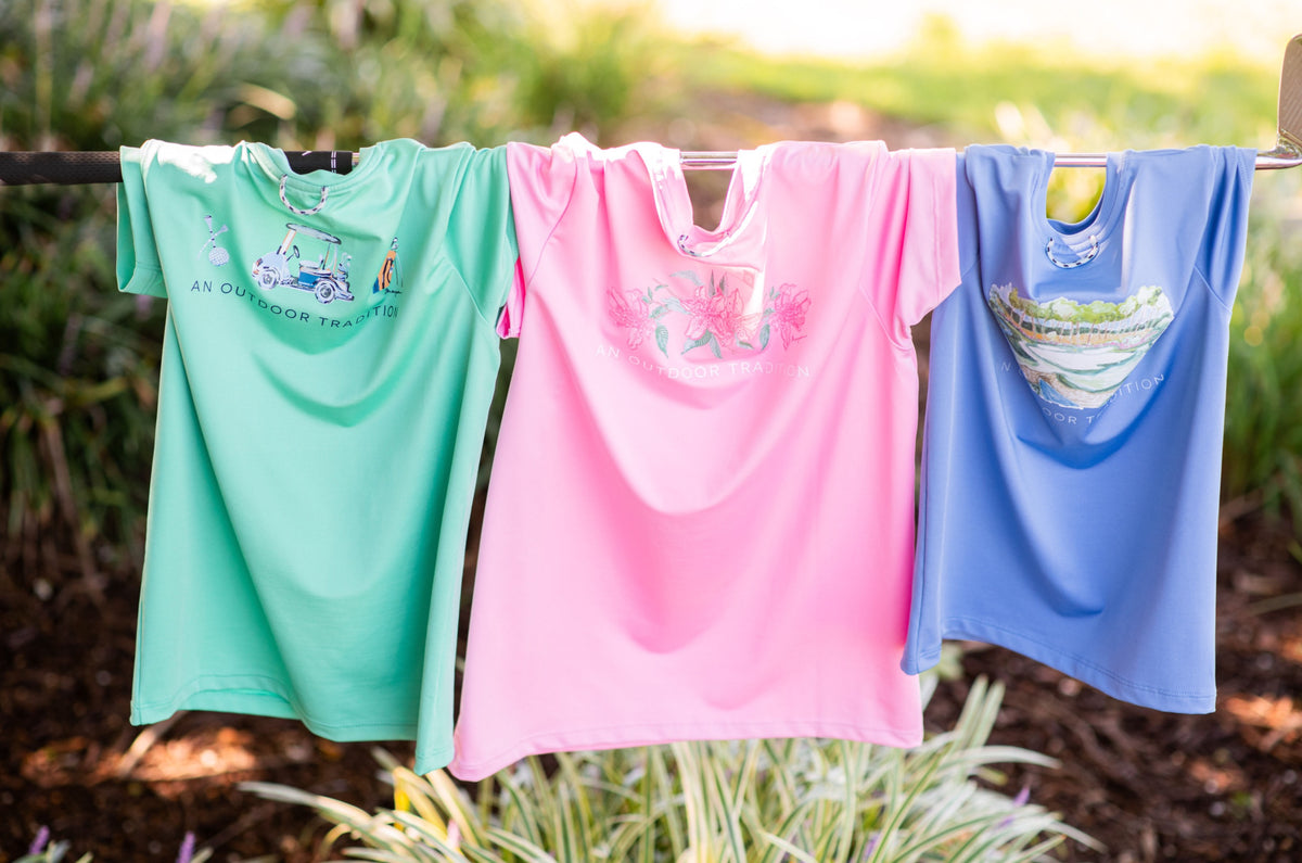 Three children's performance t-shirts in green, pink, and blue hanging on a clothesline outdoors.