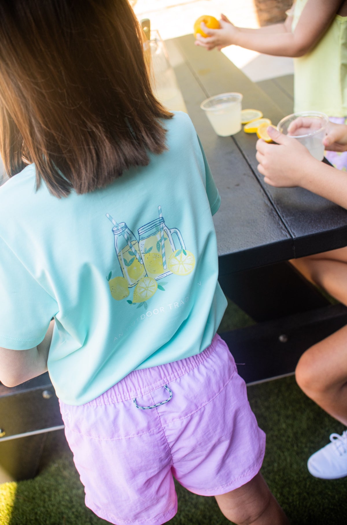 Child wearing a light blue shirt with lemon graphics and purple shorts standing near a table.
