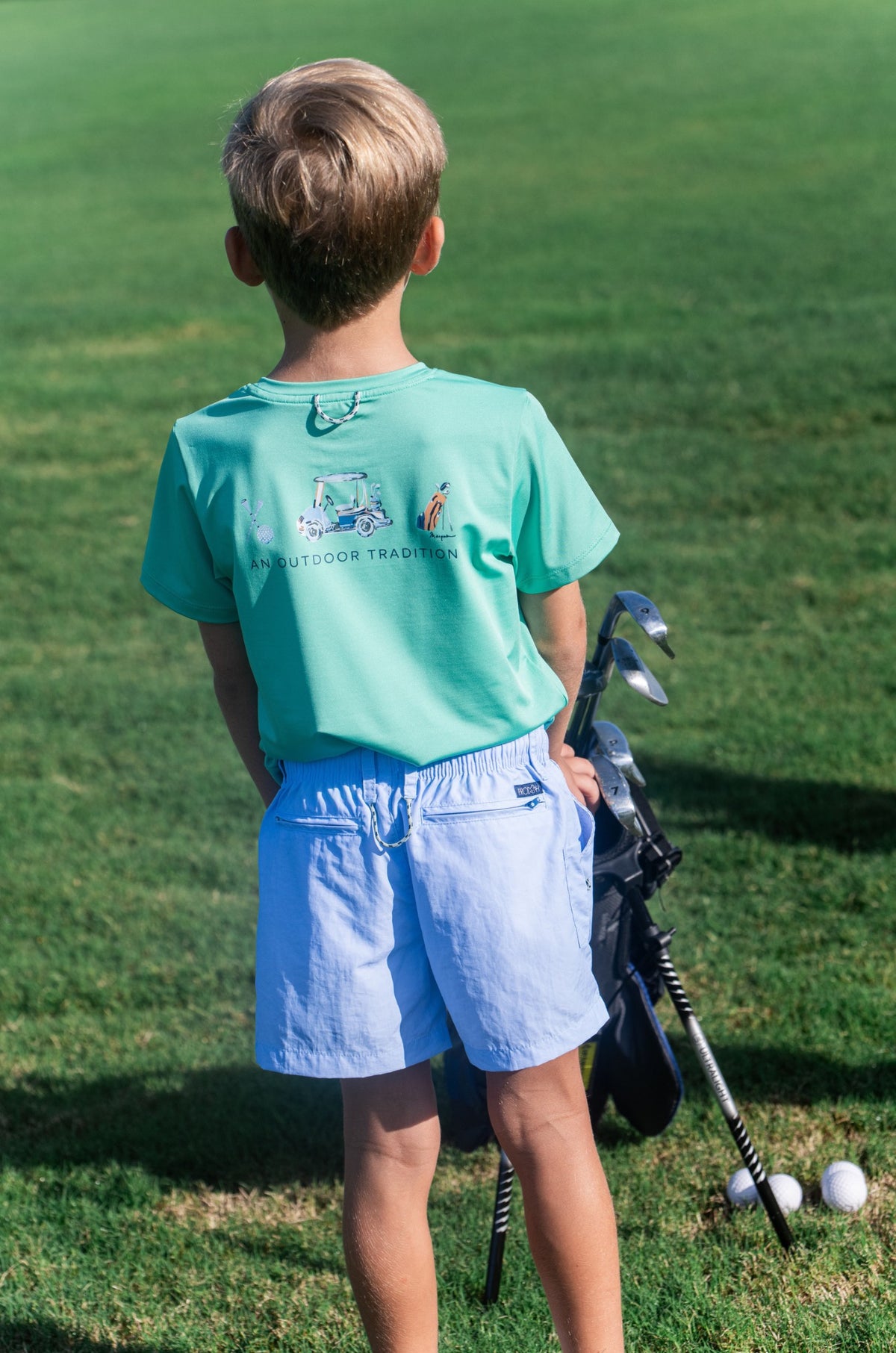Child in a green shirt and blue shorts holding golf clubs on a grassy field