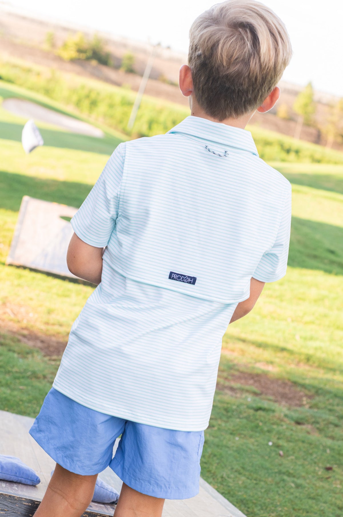 Child in light blue striped polo shirt and shorts standing on a golf course.