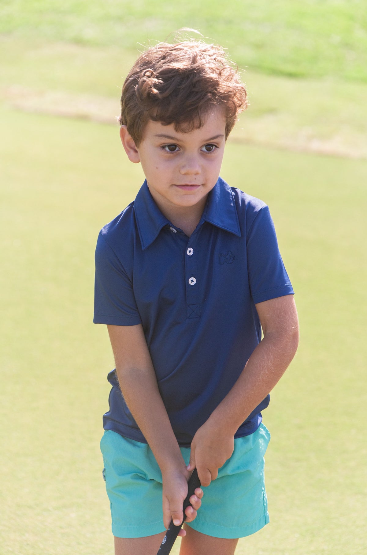 Young boy in a blue polo shirt and turquoise shorts holding a golf club on golf course