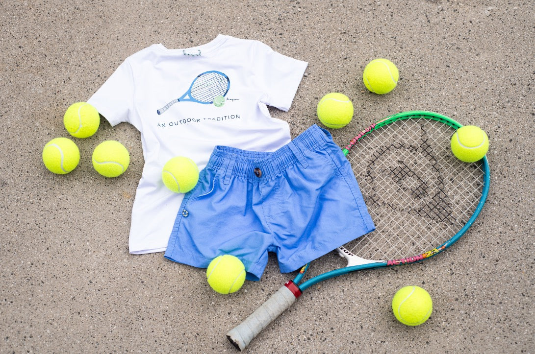 Children's tennis outfit with t-shirt, shorts, and racquet on a sandy surface with tennis balls.
