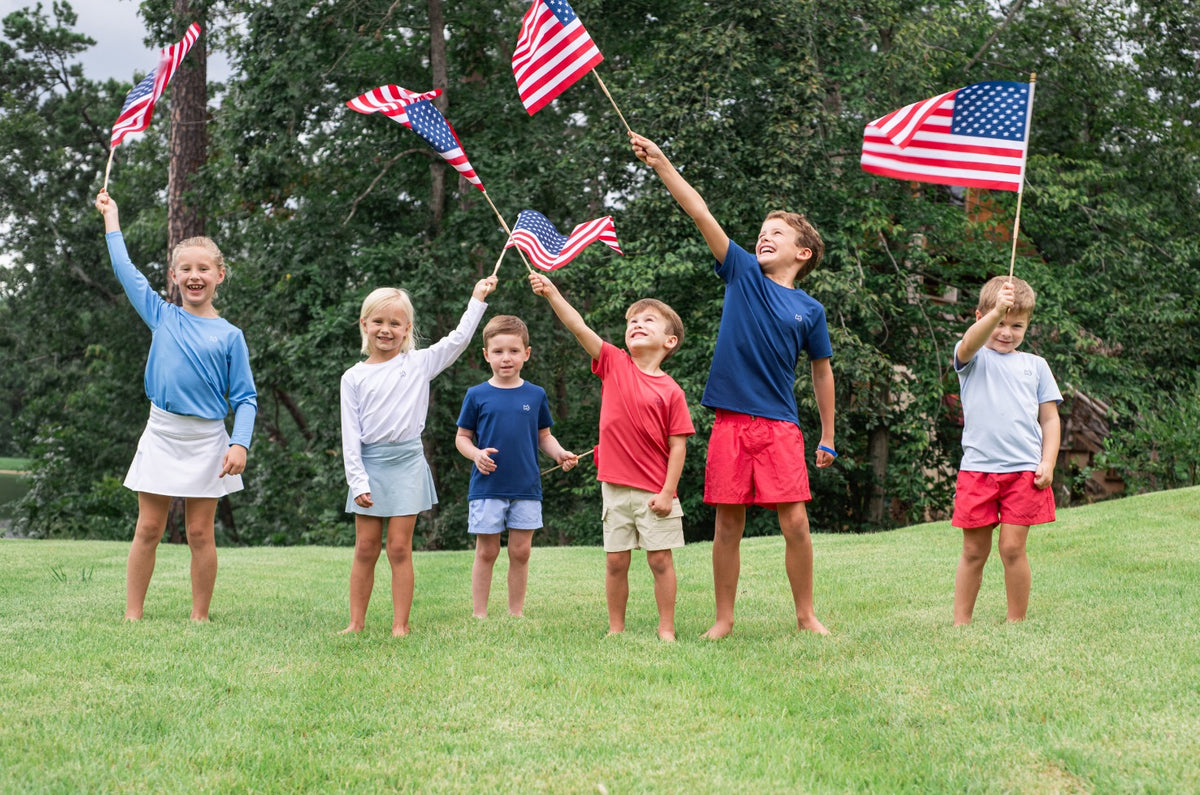 Children holding American flags on a grassy field wearing patriotic red shorts