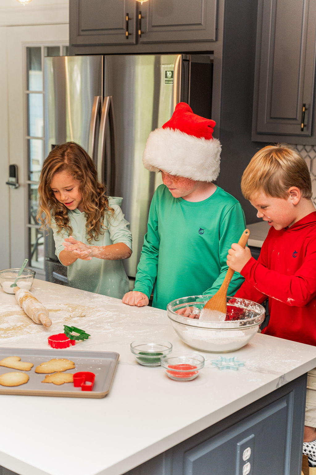 Three children baking together in a modern kitchen wearing a red Santa hat and a red long-sleeve hoodie, mixing ingredients in a large glass bowl with a wooden spoon.