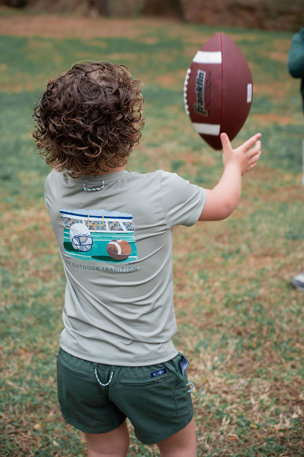 A child seen from behind wearing a light gray t-shirt with a football graphic on the back, holding a football on a grassy field.