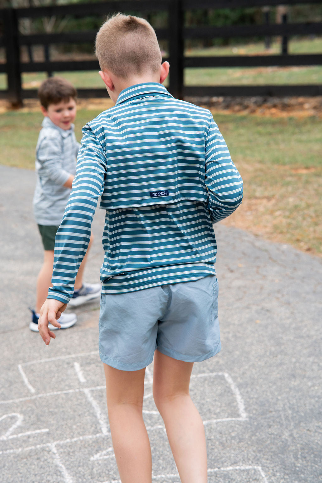 A boy seen from behind wearing a blue and teal green striped long-sleeve polo shirt and light blue shorts, playing hopscotch on an outdoor paved path.