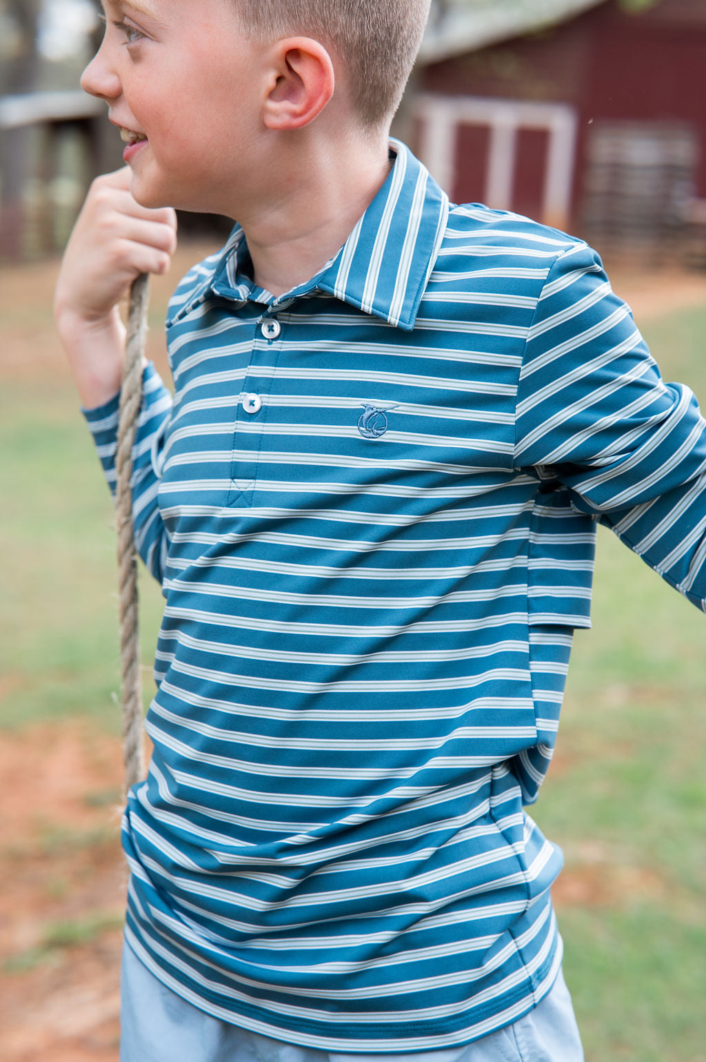 A young boy wearing a blue and white striped long-sleeve polo shirt, standing outdoors in a grassy area.