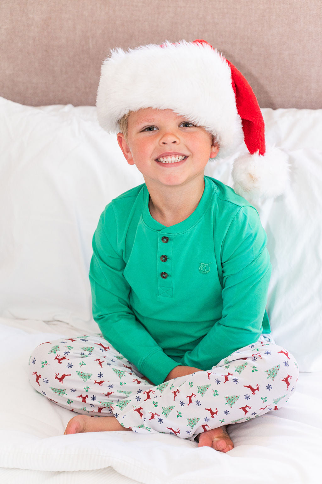 A young boy smiling while sitting cross-legged on a white bed, wearing a green long-sleeve green henley shirt, white pajama pants with a red and green Christmas tree and reindeer pattern, and a red and white Santa hat.