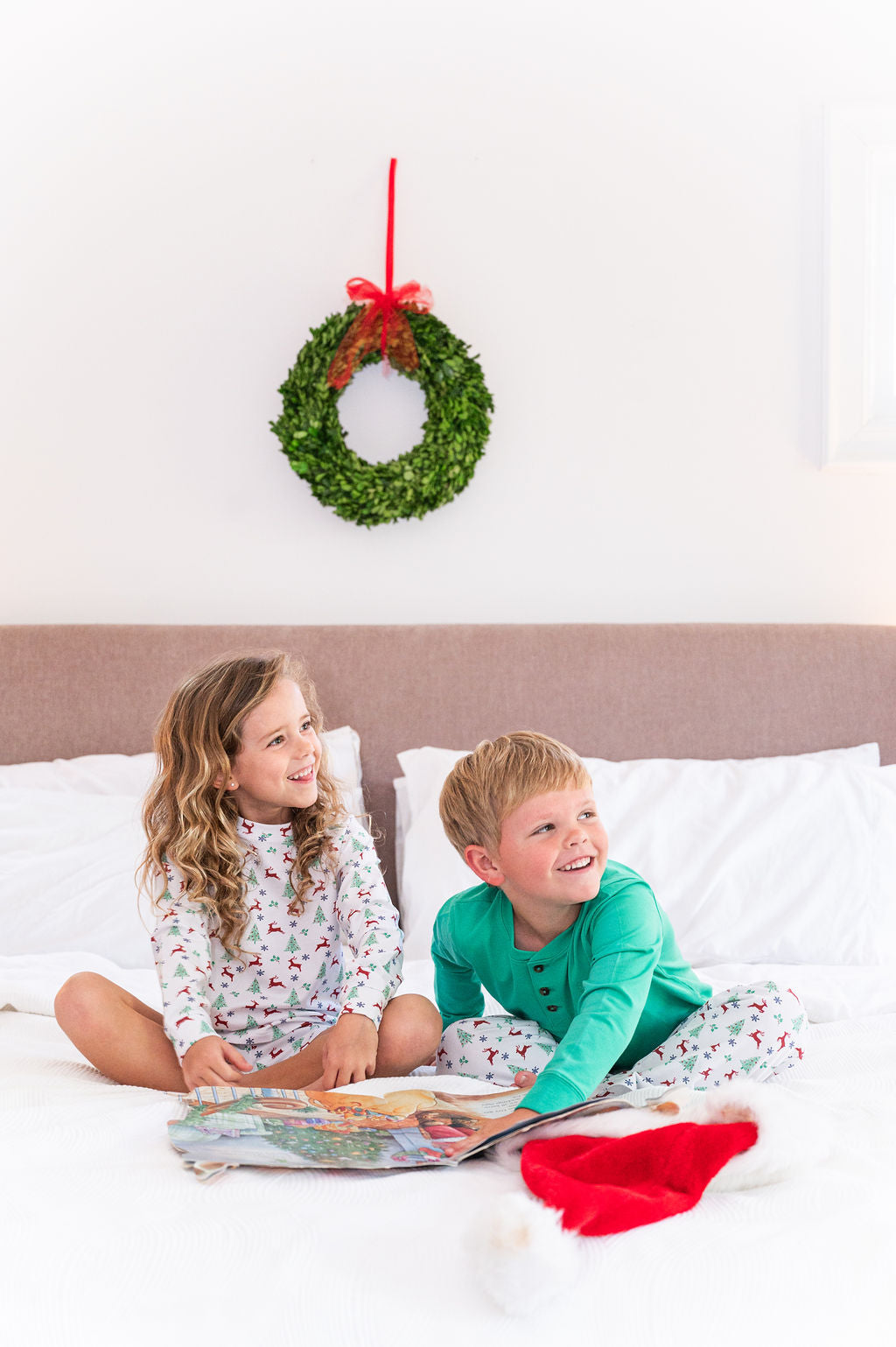 Two children sitting on a bed reading a book, with a green wreath and red bow hanging on the wall above them. A red Santa hat rests on the bed.