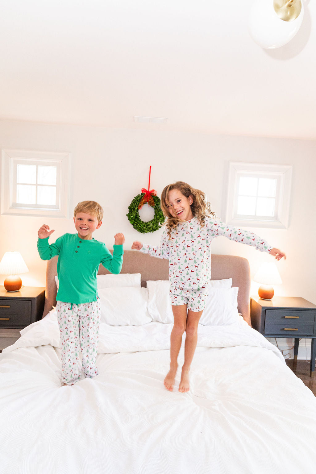Two children wearing matching holiday-themed pajamas jump on a white bed in a brightly lit bedroom. The boy wears a holly green henley long-sleeve top with white patterned pants, and the girl wears a white long-sleeve top with a small red christmas print and matching white pants.