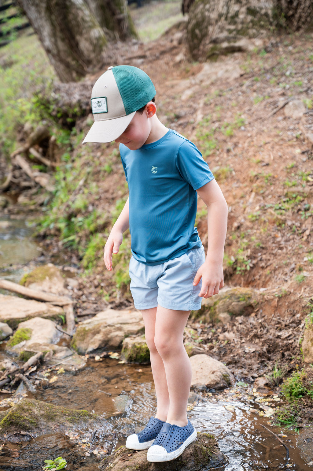 A young boy wearing a teal and beige trucker hat, a blue t-shirt fishing lure t-shirt, light blue shorts, and blue slip-on shoes stands on rocks in a shallow stream in an outdoor wooded setting.