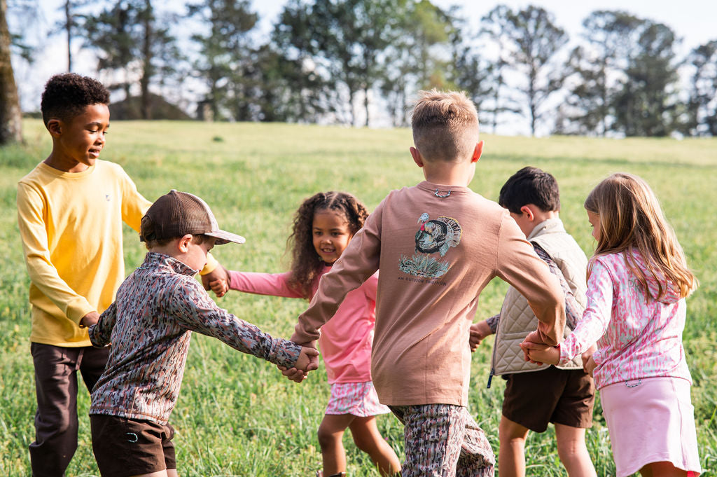 A group of children playing in a grassy park, holding hands in a circle. The children are wearing casual fall clothing, including a brown long-sleeve t-shirt with a turkey graphic
