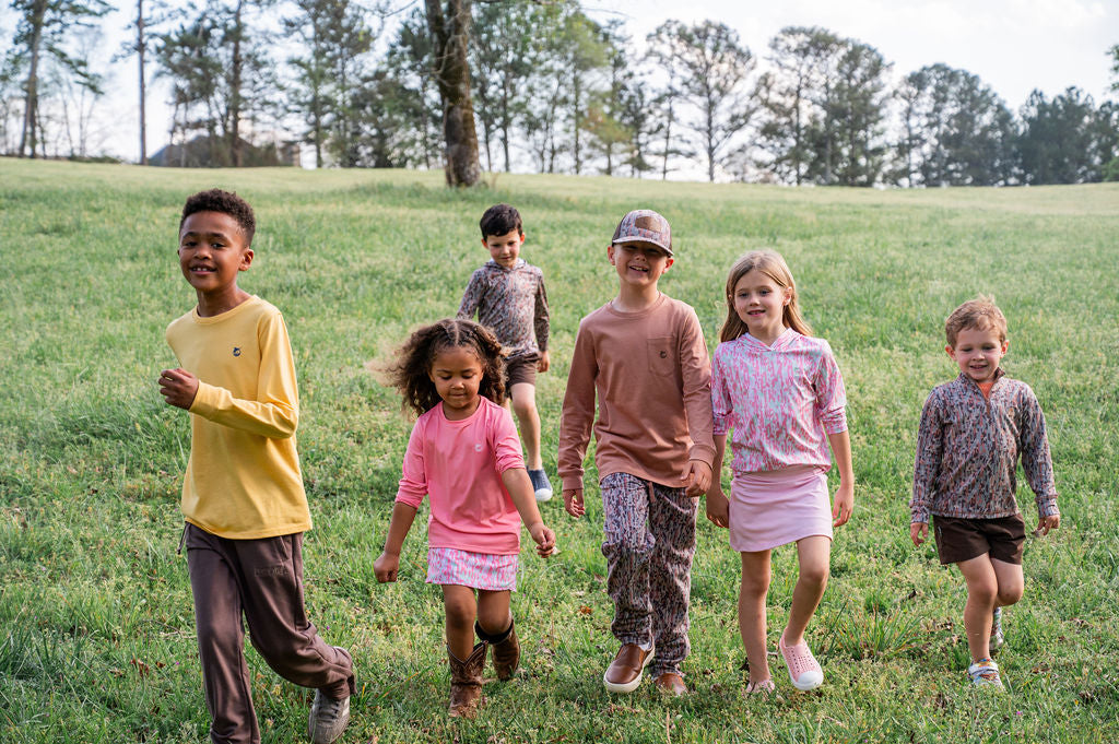 Six children walking hand-in-hand across a grassy hill outdoors, wearing casual, coordinated outfits in shades of yellow, pink, brown, and gray.