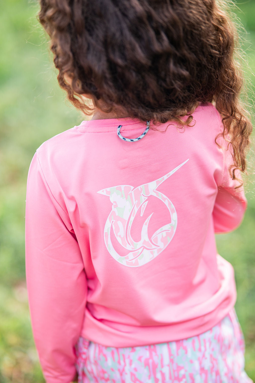 A child viewed from behind wearing a bright pink long-sleeve t-shirt with a large white fish graphic on the back.
